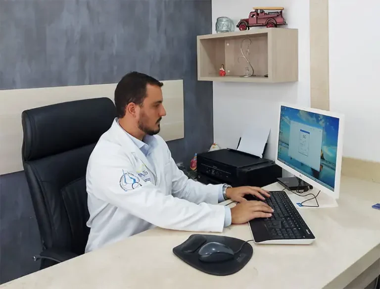 Dr. Dieno Portella, especialista em ortopedia, trabalhando em seu consultório no computador. Ele está sentado em uma cadeira preta, vestindo um jaleco branco com o logotipo do Instituto Nacional de Ortopedia e Traumatologia (INRO) bordado. O consultório tem uma parede cinza e uma estante com decorações.