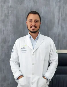 Dr. Dieno Portella, especialista em ortopedia, em pé no consultório, vestindo um jaleco branco com o logotipo do Instituto Nacional de Ortopedia e Traumatologia (INRO) bordado. Ele está sorrindo e tem as mãos nos bolsos do jaleco. A parede de fundo é cinza.