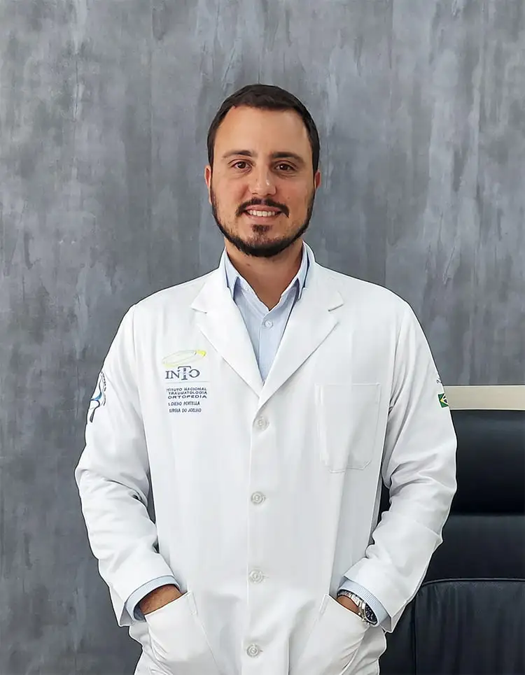 Dr. Dieno Portella, especialista em ortopedia, em pé no consultório, vestindo um jaleco branco com o logotipo do Instituto Nacional de Ortopedia e Traumatologia (INRO) bordado. Ele está sorrindo e tem as mãos nos bolsos do jaleco. A parede de fundo é cinza.